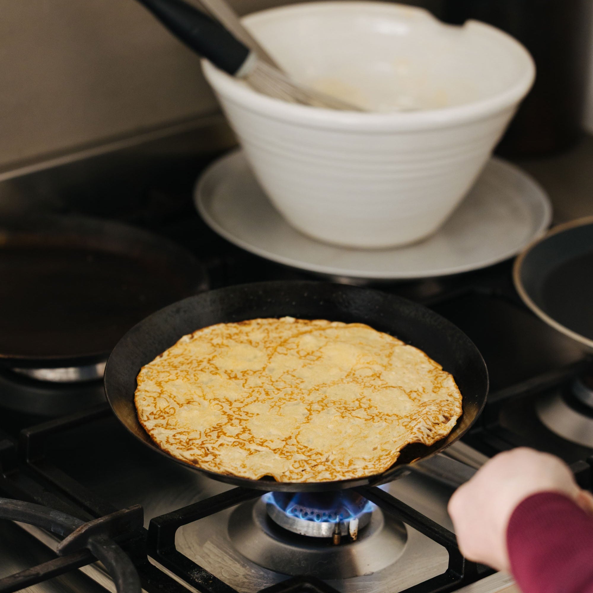 Person cooking a pancake on a gas stove with a bowl and spoon in the background.
