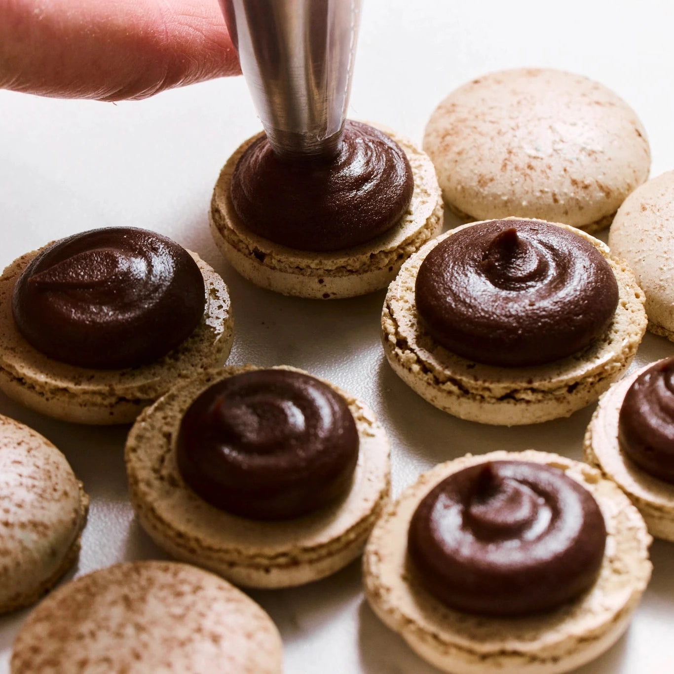 Person filling macarons with chocolate ganache on a white surface